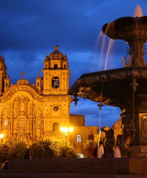 Arquitectura de estilo colonial de la Catedral del Cusco iluminada de noche, con una fuente decorativa en primer plano y un cielo dramático.