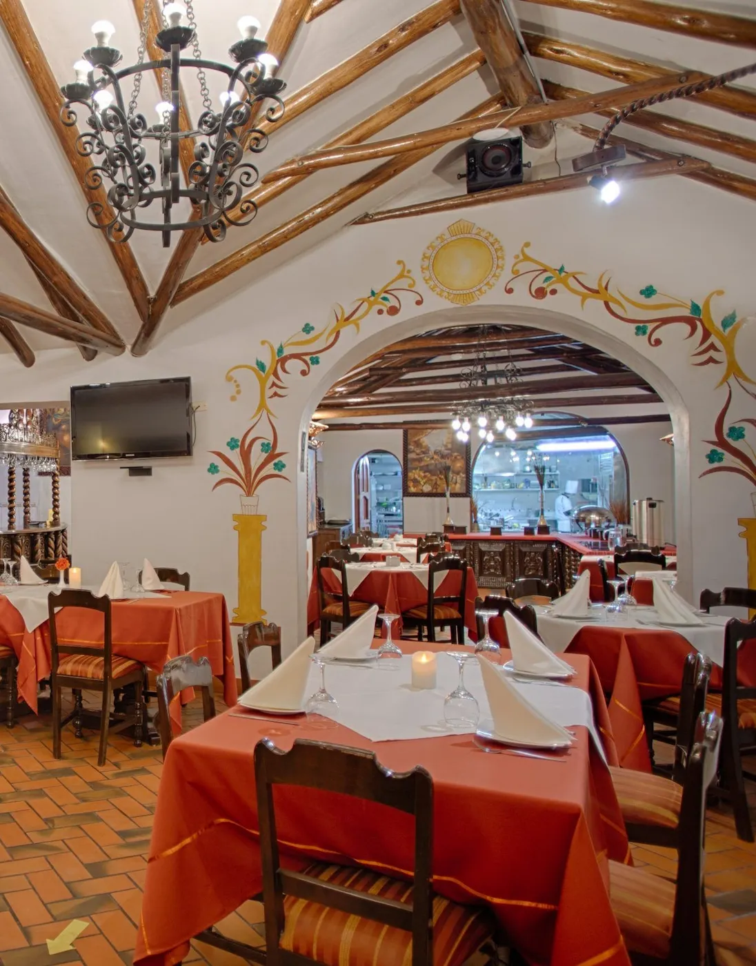 Vista interior del restaurante de Munay Wasi Inn con decoración de estilo colonial, vigas de madera, arte mural vibrante y mesas con manteles naranjas, servilletas blancas y velas, destacando el ambiente acogedor para cenar en Cusco, Perú.